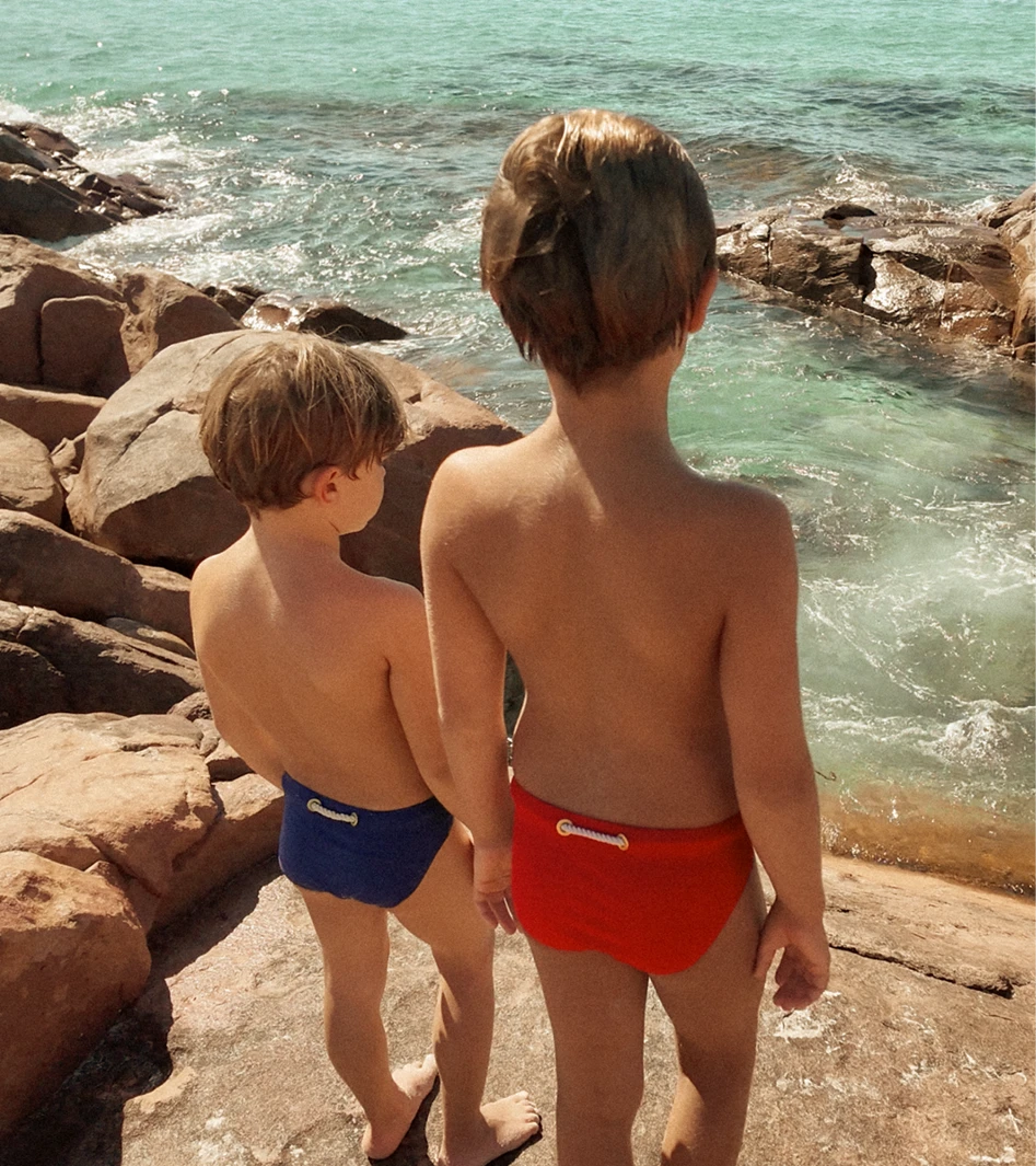 Two children wearing red and navy Lobby’s Nippers, standing on a rocky Australian shoreline overlooking the ocean.