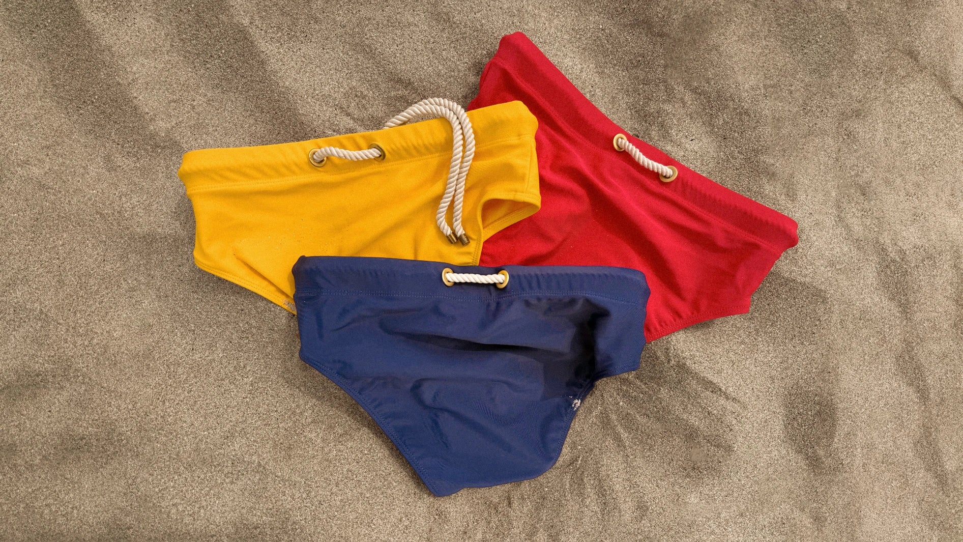 Lobby’s Nippers yellow, red and navy swim briefs arranged on sand, featuring signature rope drawstring details - designed for children. 